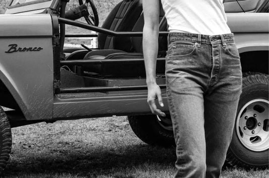 Heavyweight denim jeans paired with a white tank top beside a vintage Bronco vehicle.