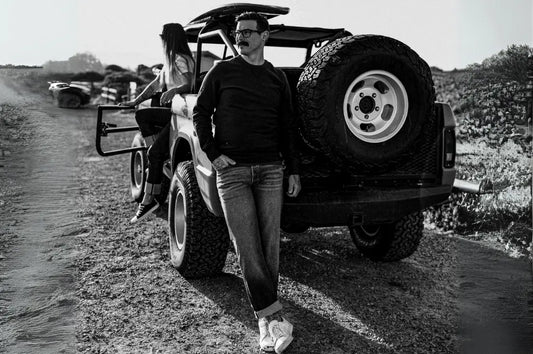 Raw denim jeans worn by man leaning against off-road vehicle in rugged outdoor setting with black and white filter