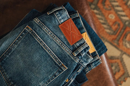 Unwashed denim jeans stacked on a leather surface, featuring contrast stitching and leather brand patches