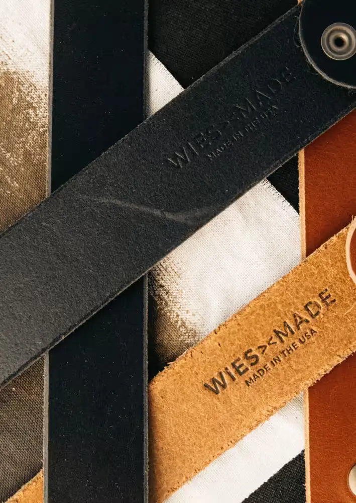 Latigo Belt in black 8oz American Latigo Leather, 1.7” width, with embossed branding, displayed on a mixed fabric background