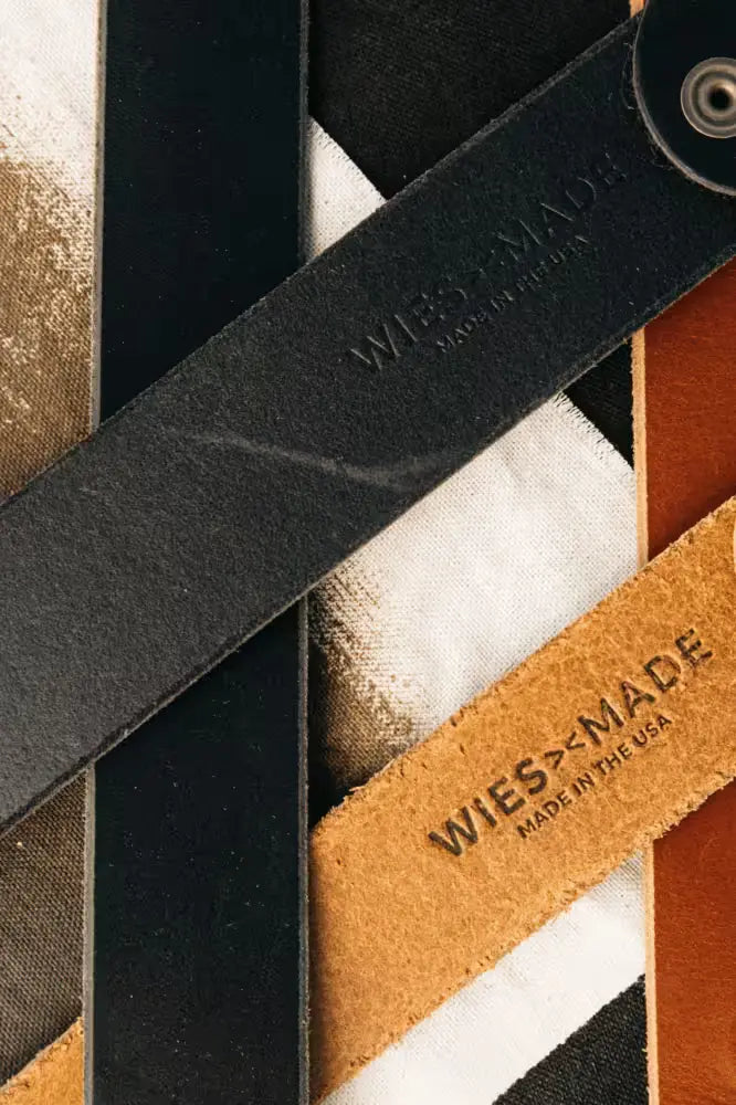 Latigo Belt in black 8oz American Latigo Leather, 1.7” width, with embossed branding, displayed on a mixed fabric background