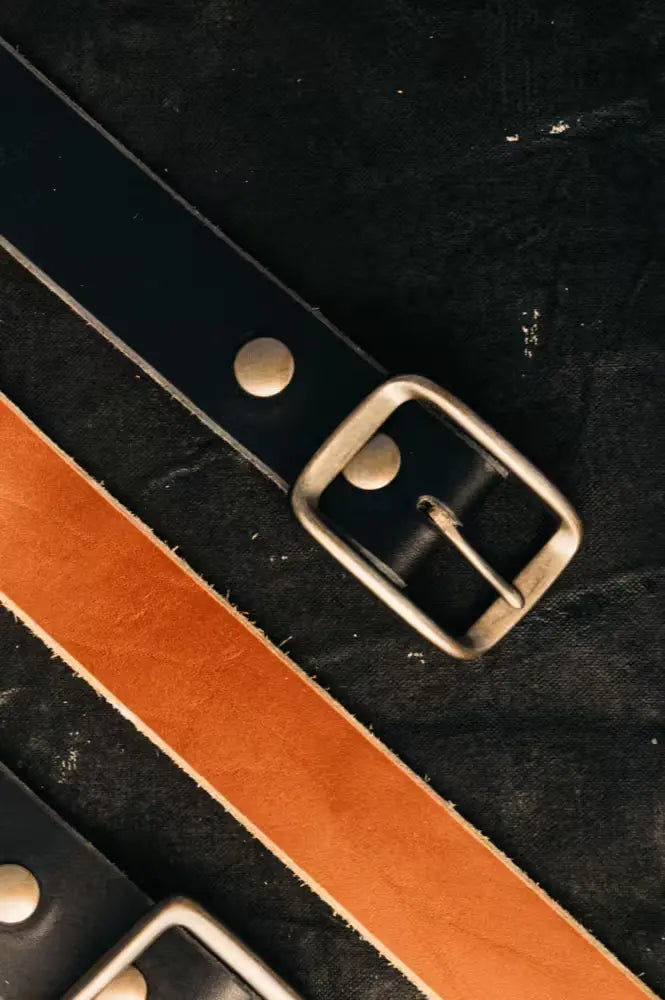 Two Latigo Belts in 1.5” width, crafted from 8oz American Latigo Leather, one in black & one in saddle color, with solid brass buckles on black fabric