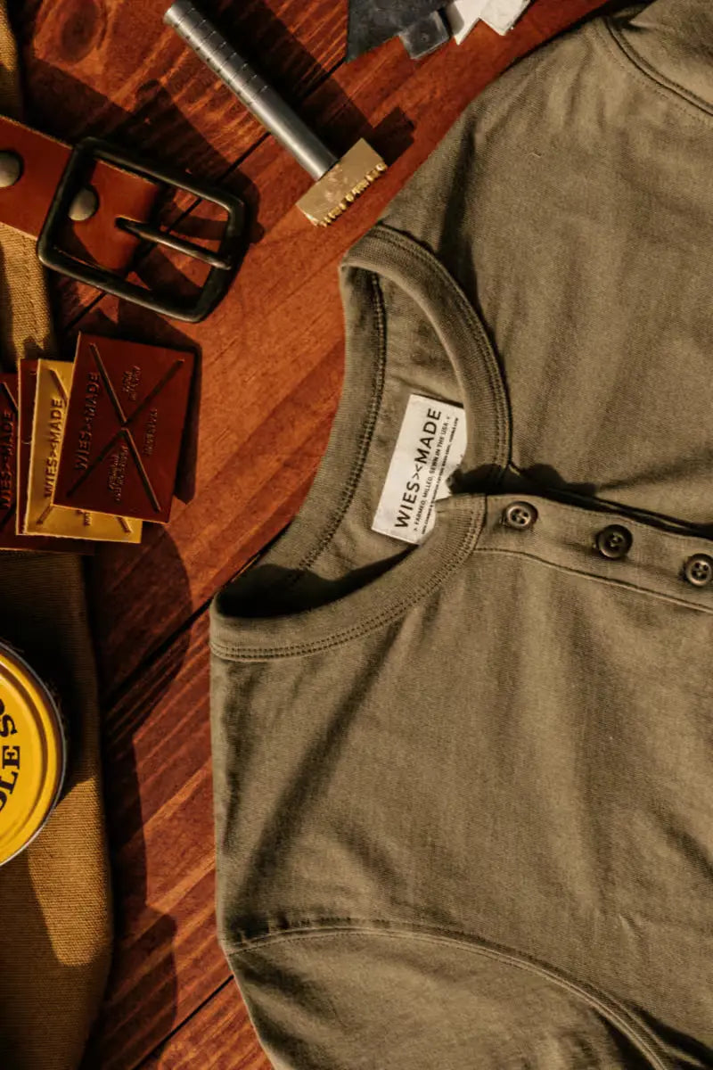 Reyes Henley in heavyweight 8oz 100% cotton olive color displayed on a wooden surface with accessories