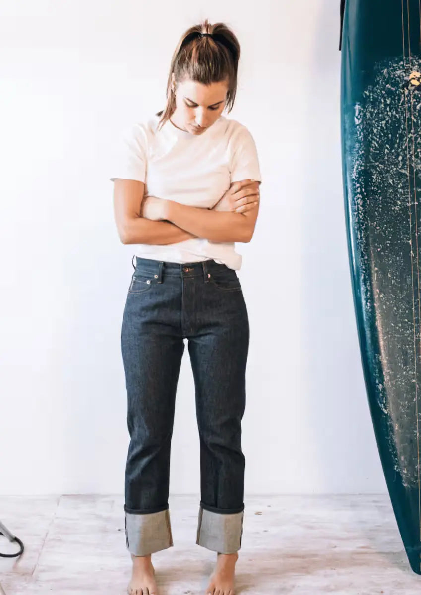 Woman with a ponytail wearing a white T-shirt and Sidney Fit Jean in raw selvedge denim, standing barefoot with arms crossed near a surfboard in a bright, minimalist room
