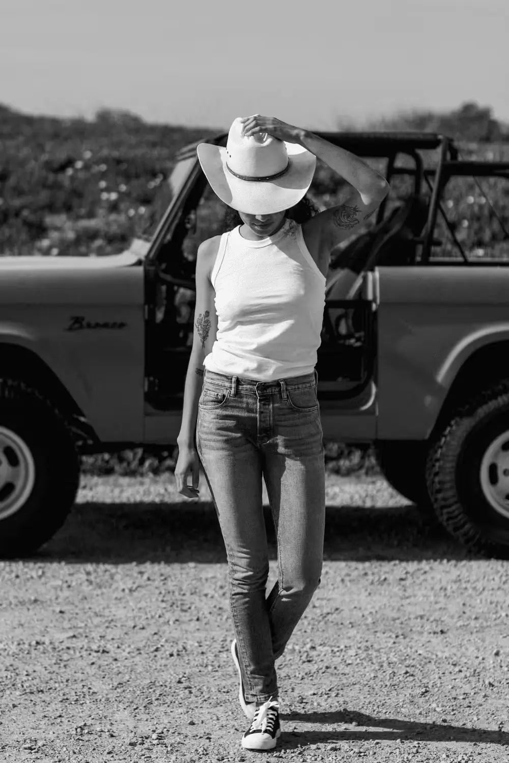 Elise Fit Jean in Salt Water Wash color, mid-rise slim fit, worn by a woman in a white tank top & cowboy hat, standing in front of a vintage off-road vehicle on a gravel surface