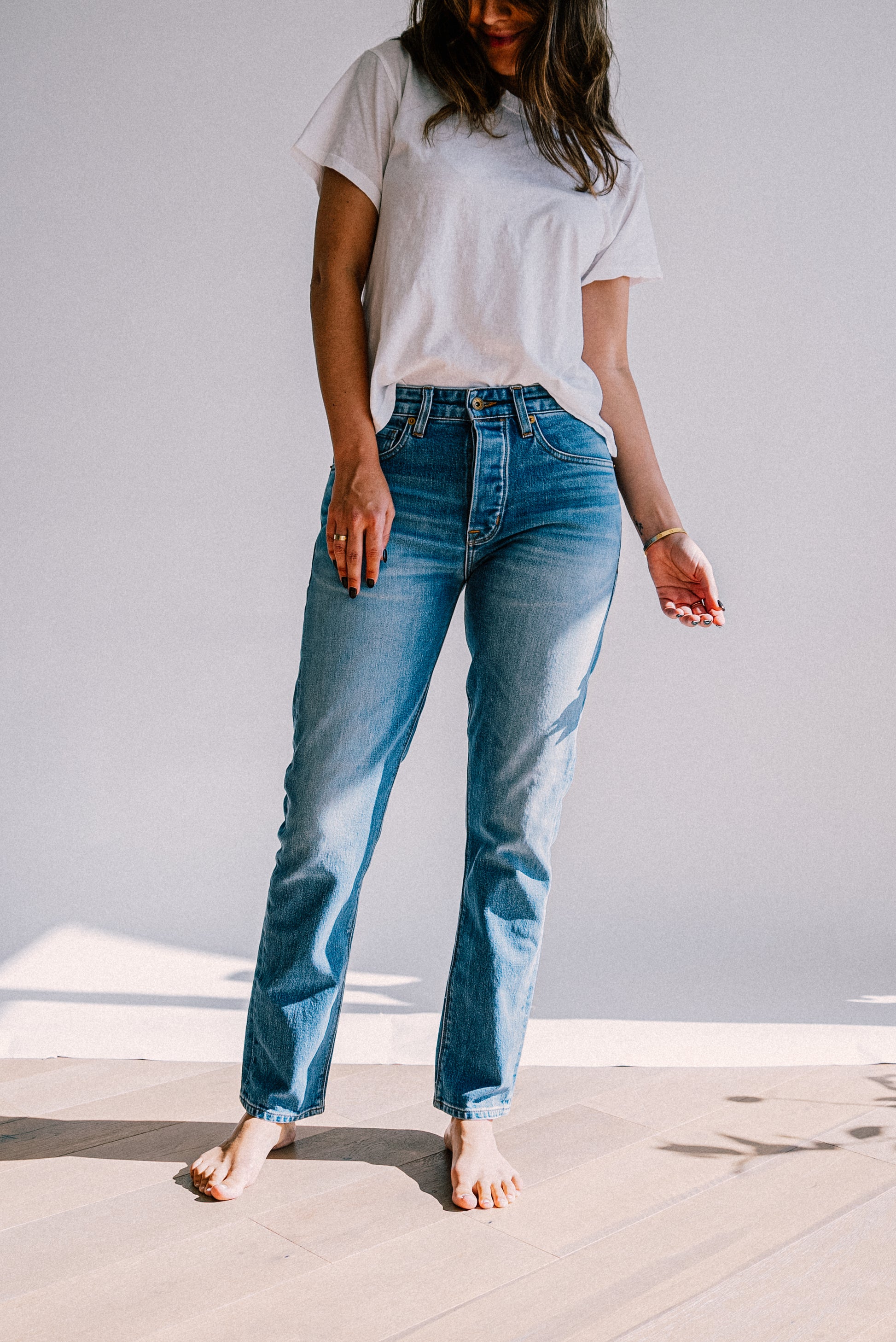 Women's denim jeans styled with a white t-shirt and worn barefoot on a sunlit wooden floor.
