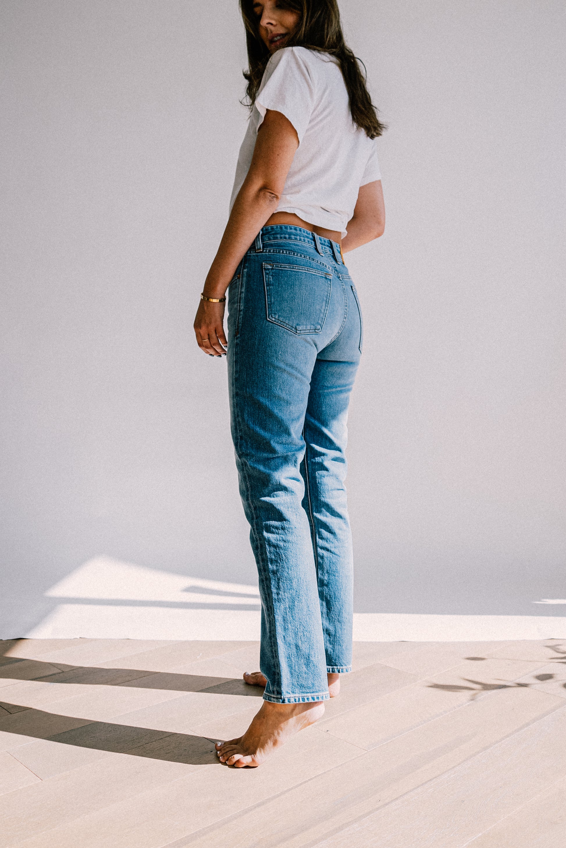 Women's tailored jeans paired with a white t-shirt, shown from the back in natural sunlight on a wooden floor.