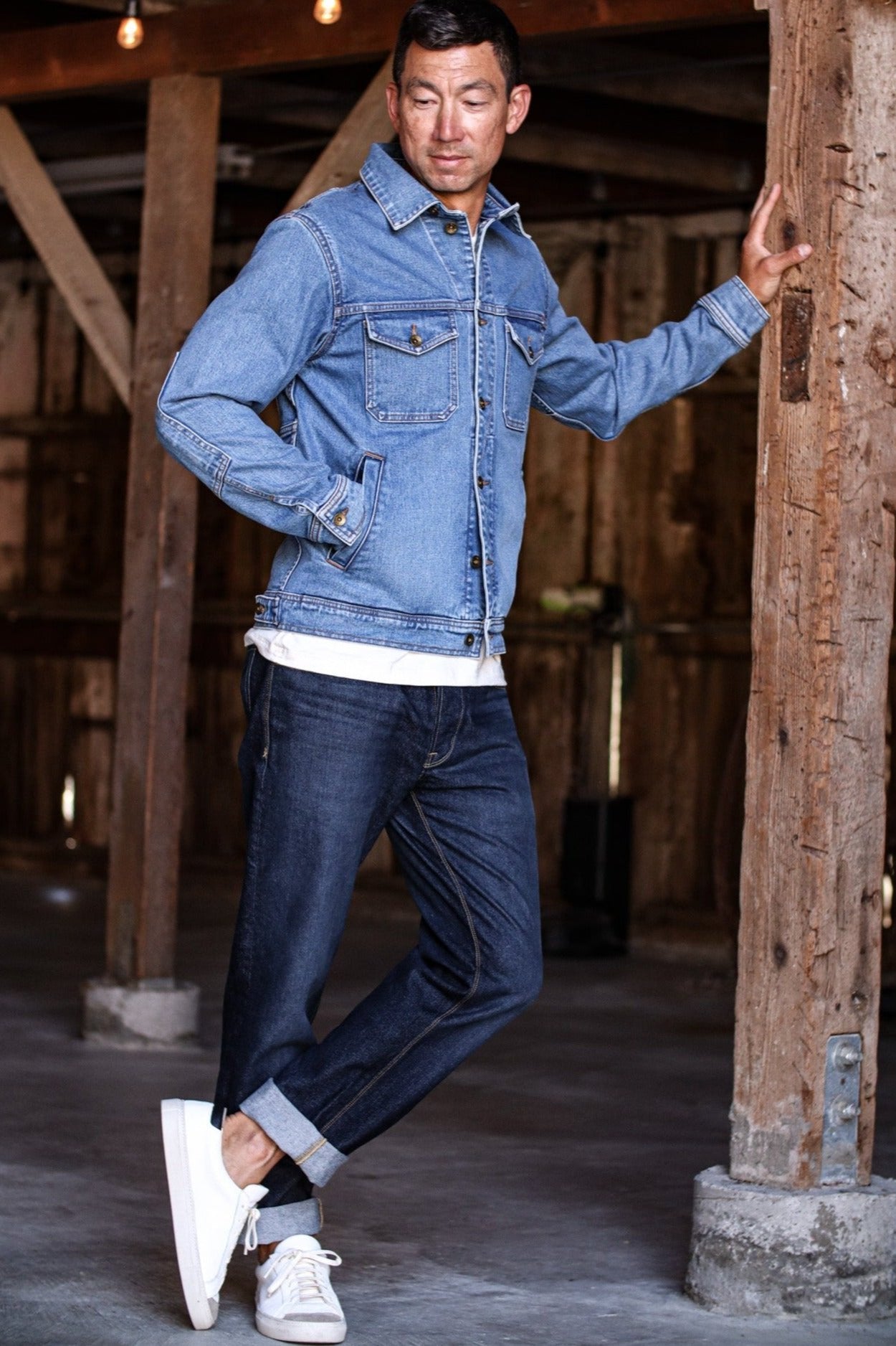 Denim jacket made in USA paired with dark jeans and white sneakers in a rustic barn setting.