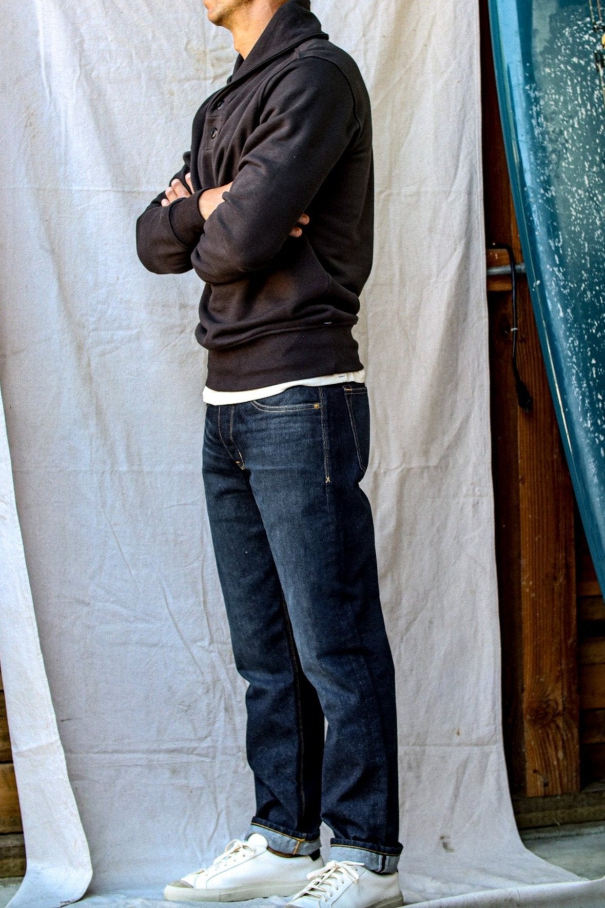 Men's shawl cardigan in black, styled with cuffed jeans and white sneakers, captured against a simple fabric backdrop.