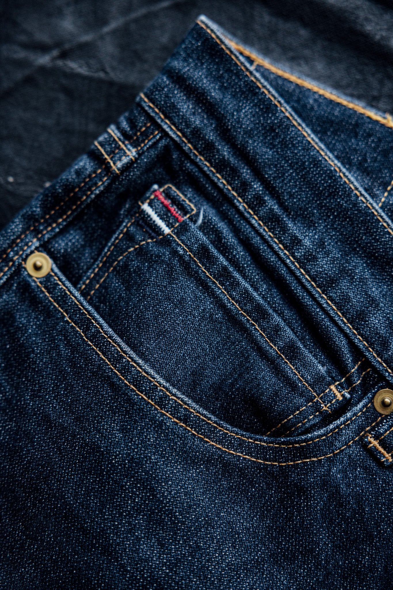 Real denim jeans with a close-up view of the pocket, featuring contrast stitching, brass rivets, and selvedge detailing.