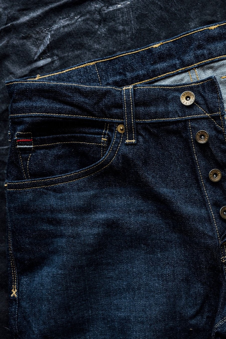 Dark American denim jeans with contrast stitching, button fly, and selvedge detailing, laid flat on a textured black background.