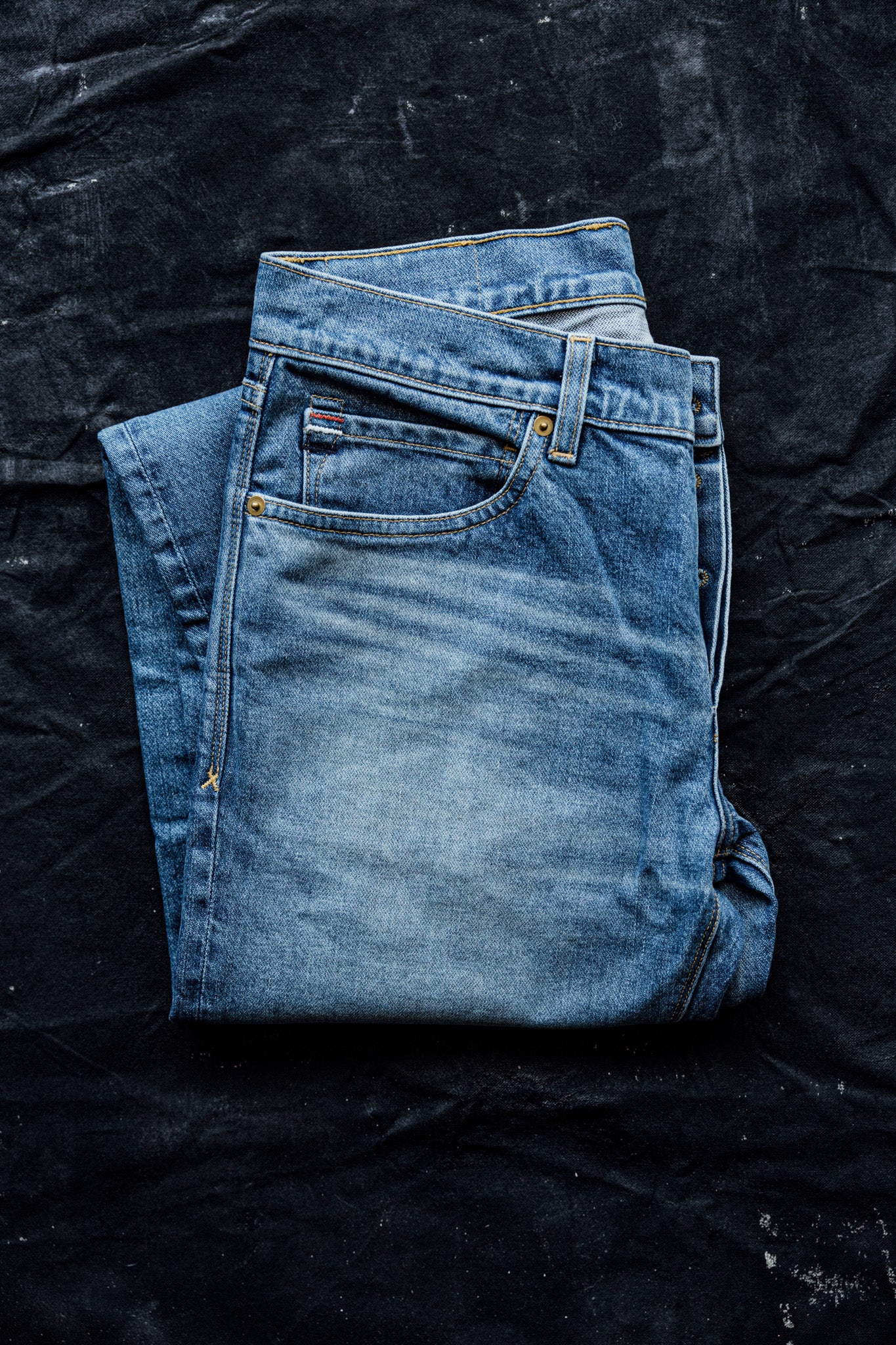Men's selvedge denim jeans in a light wash with natural fading, folded to showcase stitching and coin pocket details on a black fabric background.