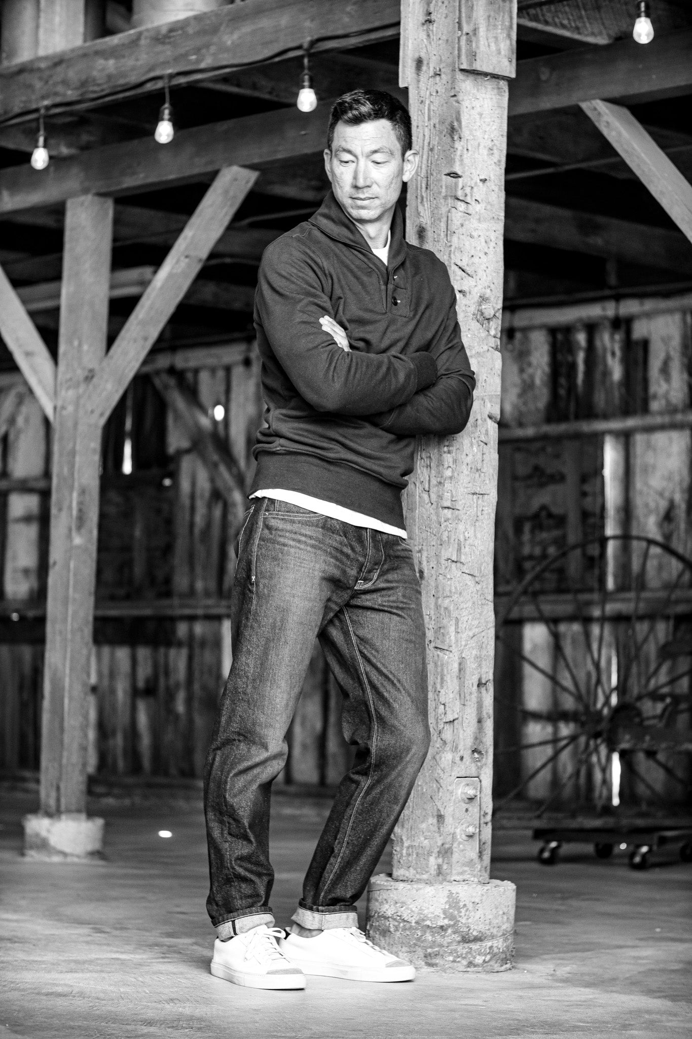 Men's raw selvedge denim jeans styled with a dark pullover, white t-shirt, and white sneakers in a rustic setting.