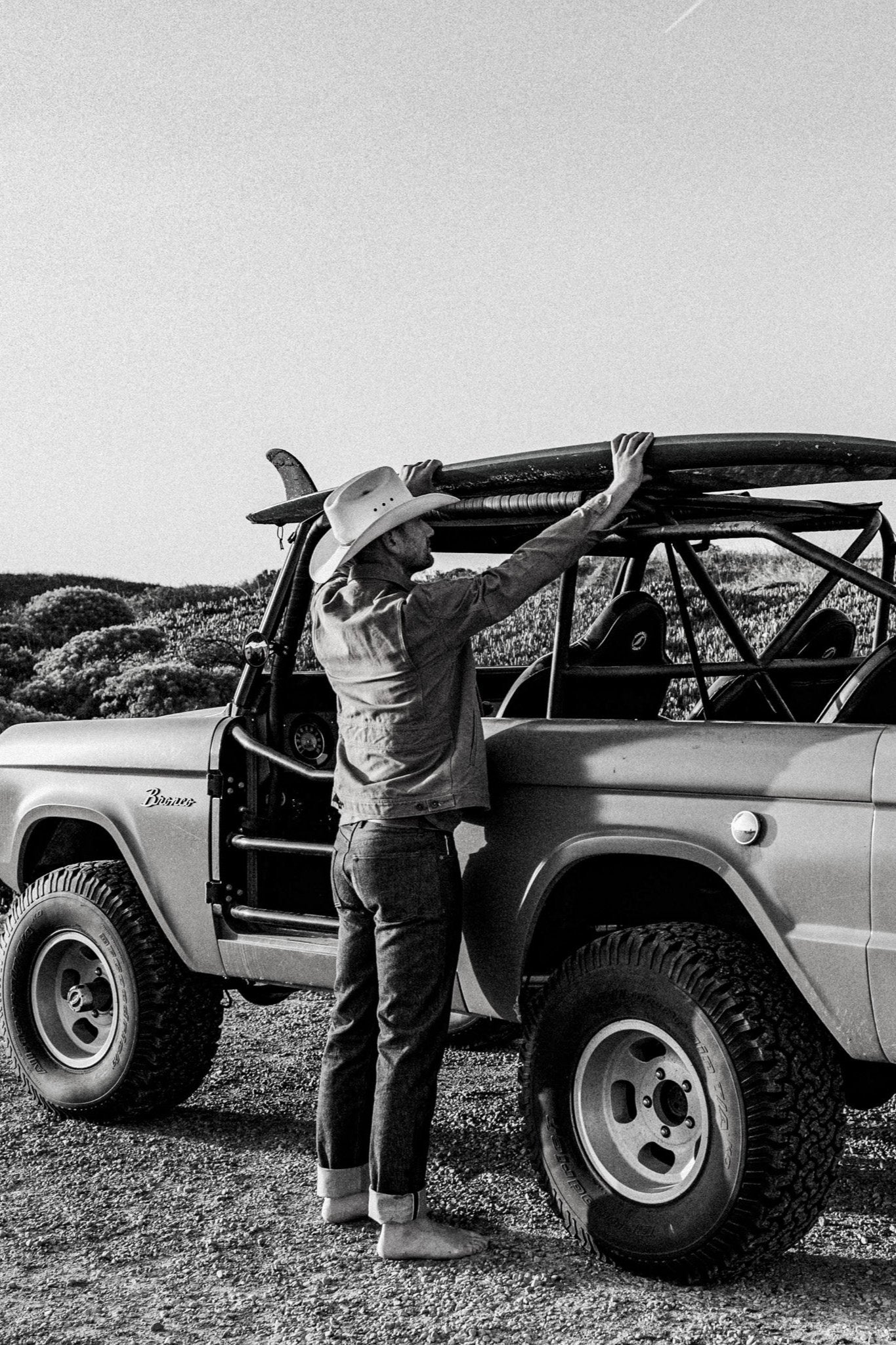 Black raw denim jeans with cuffed hems worn by a man in a cowboy hat, loading a surfboard onto a vintage Bronco.
