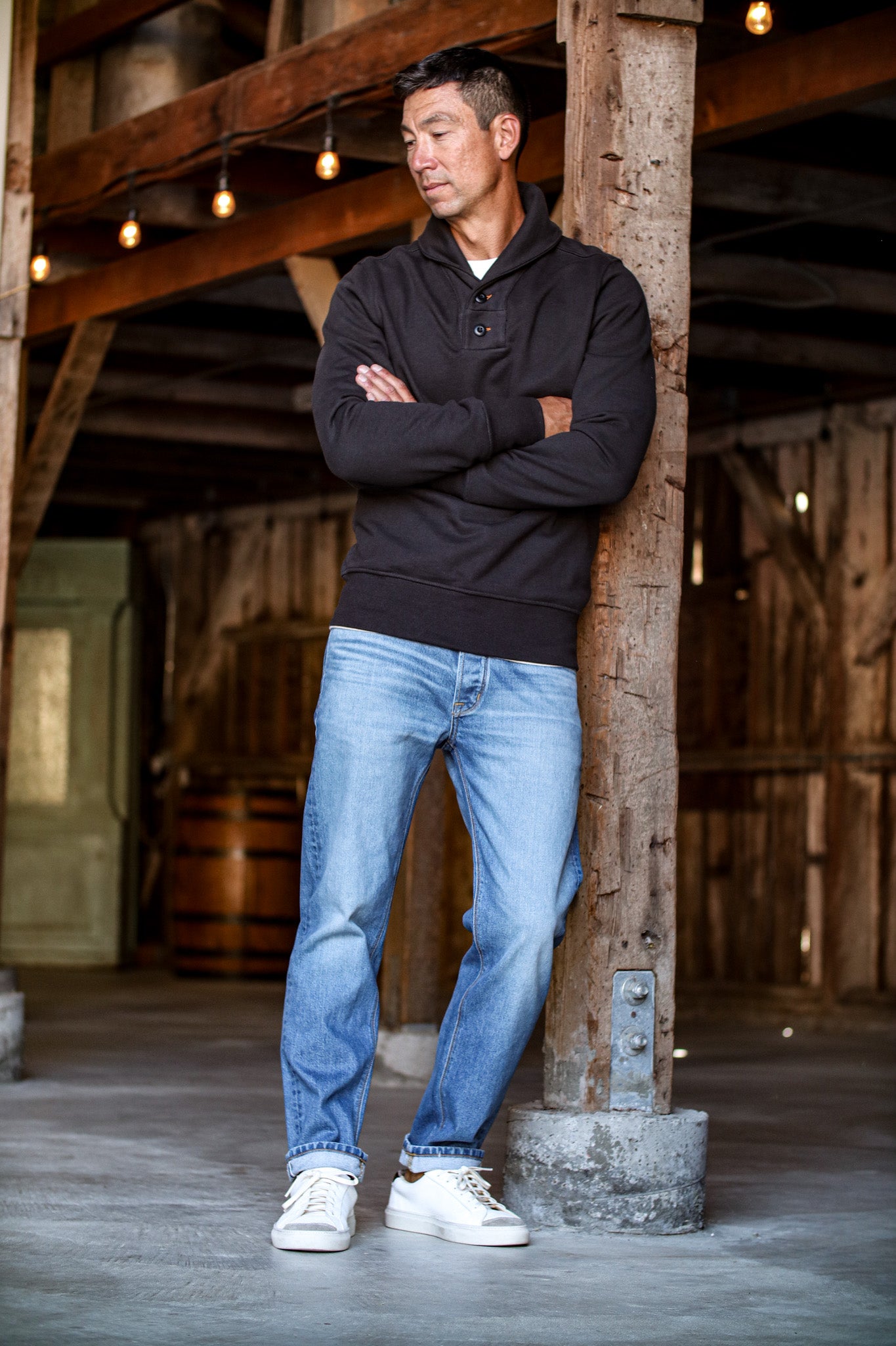 Raw selvedge denim jeans for men in a light wash with natural fading, styled with a black pullover and white sneakers in a rustic setting.