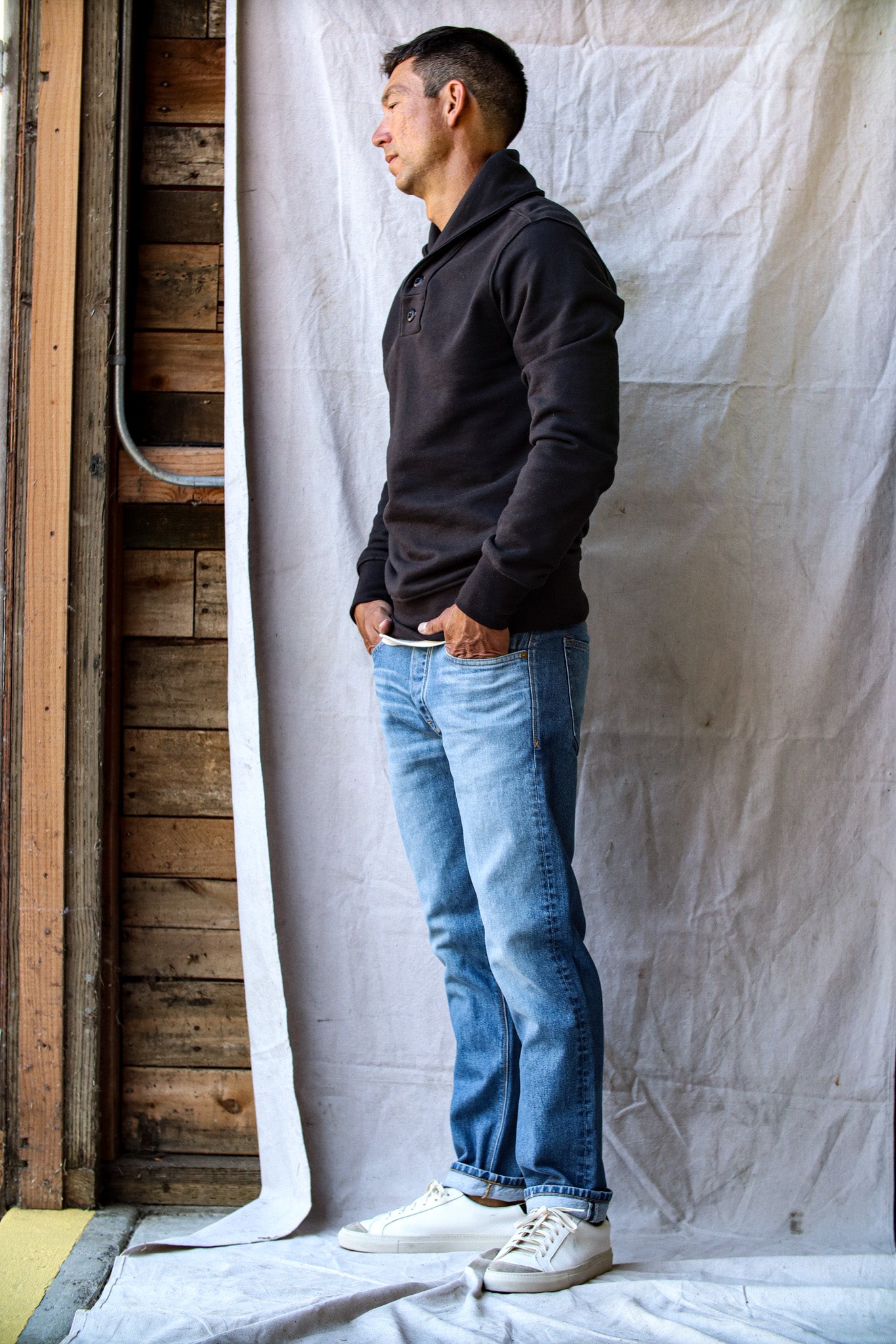 Men's natural denim jeans in a light wash with subtle fading, styled with a black pullover and white sneakers against a rustic backdrop.