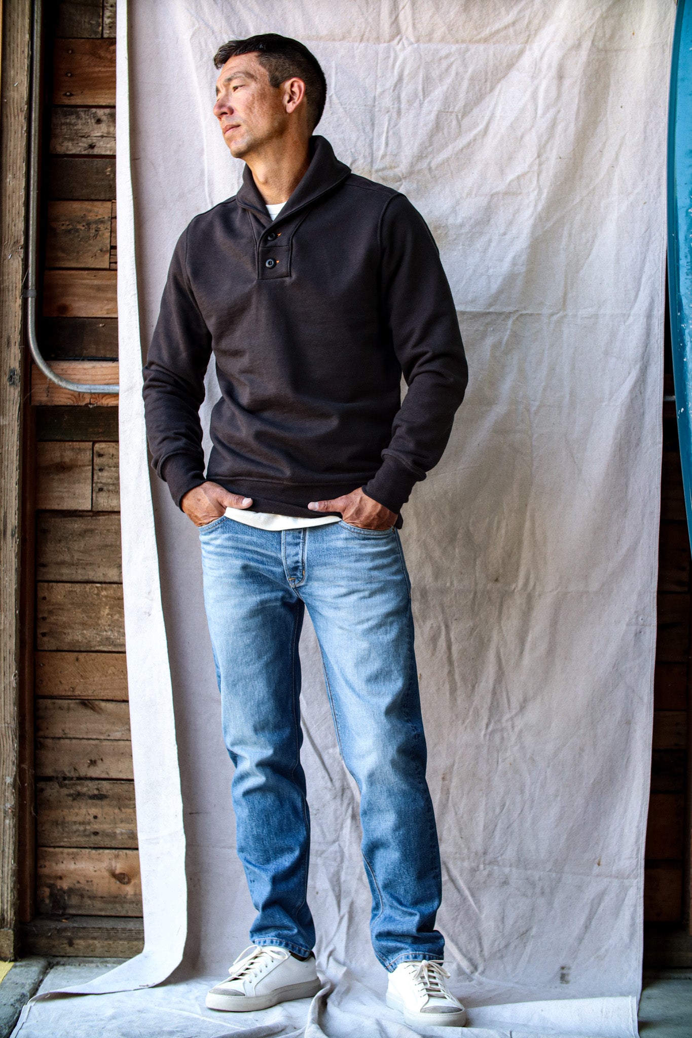 Men's selvedge denim jeans in a light wash with natural fading, styled with a black pullover and white sneakers against a rustic backdrop.