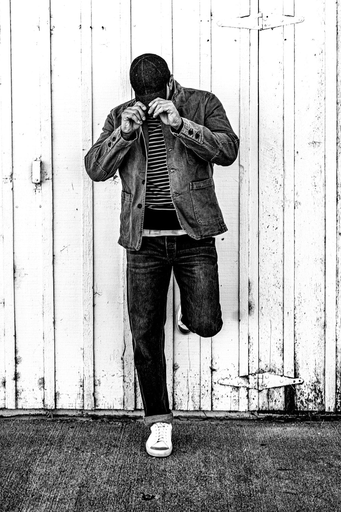 Black and white photo of a man adjusting his collar in a men's wheat denim chore blazer, one leg raised against a barn door