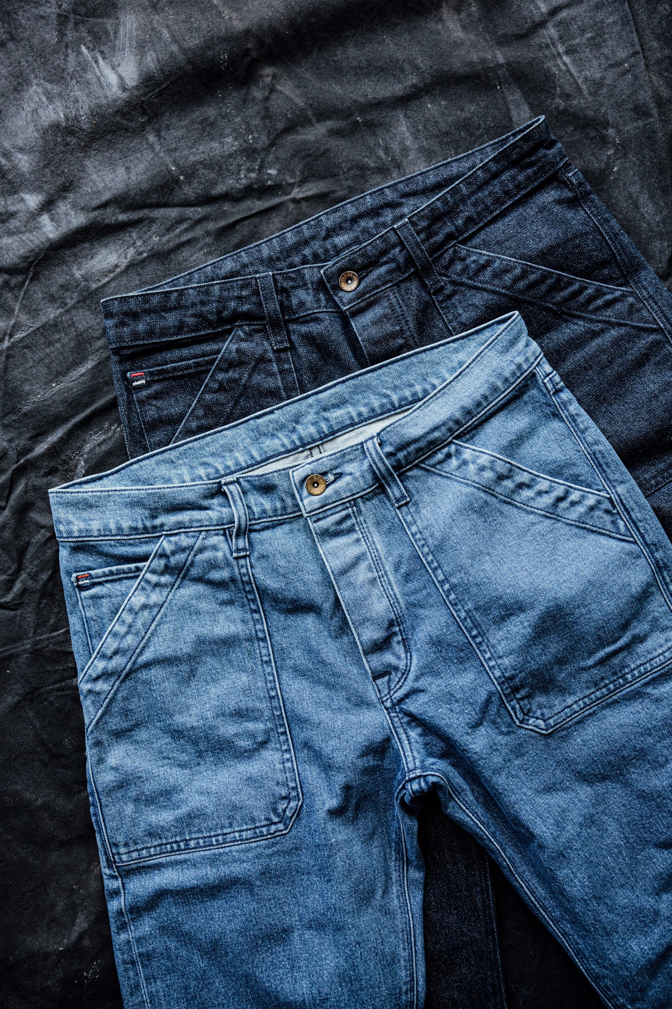 Two pairs of selvedge denim jeans, one light wash and one dark wash, laid flat on a textured black surface.