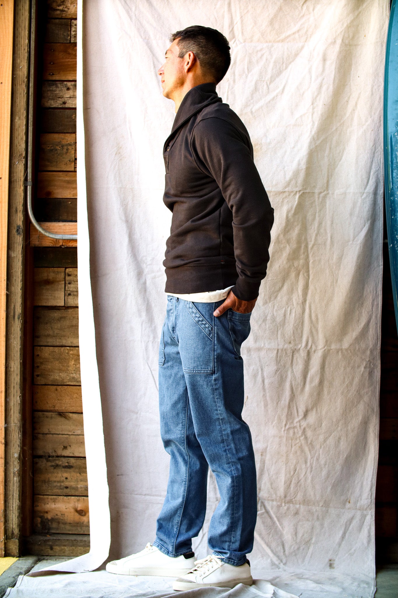 Men's raw denim jeans in a relaxed fit, styled with a black pullover and white sneakers against a rustic backdrop.