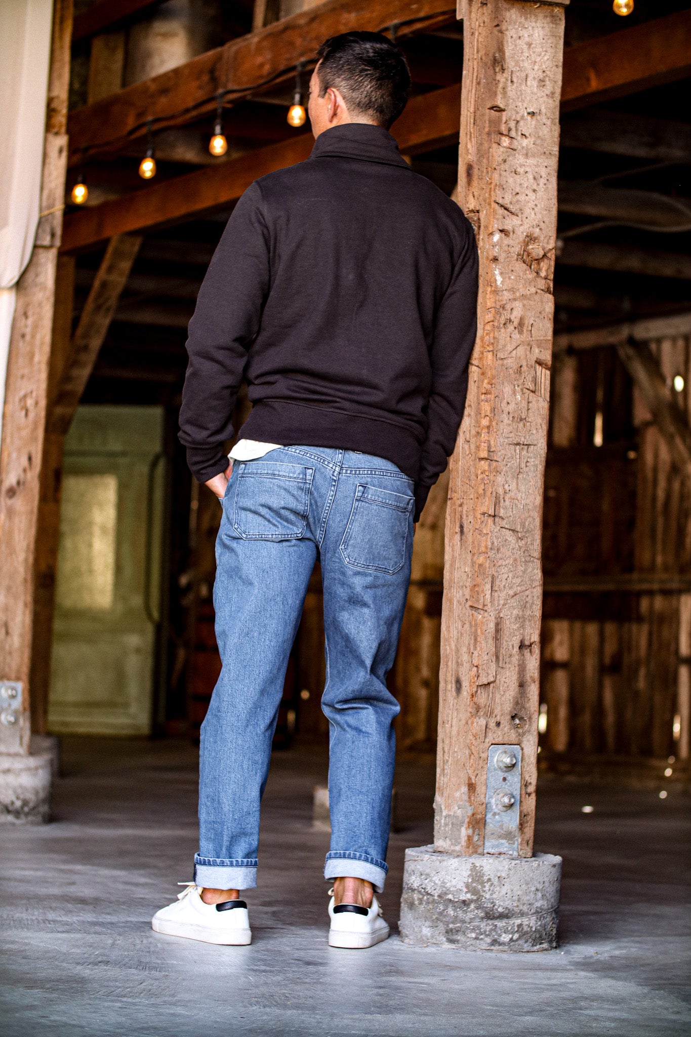 Men's selvedge denim jeans in a relaxed fit, styled with a black sweatshirt and white sneakers in a rustic setting.