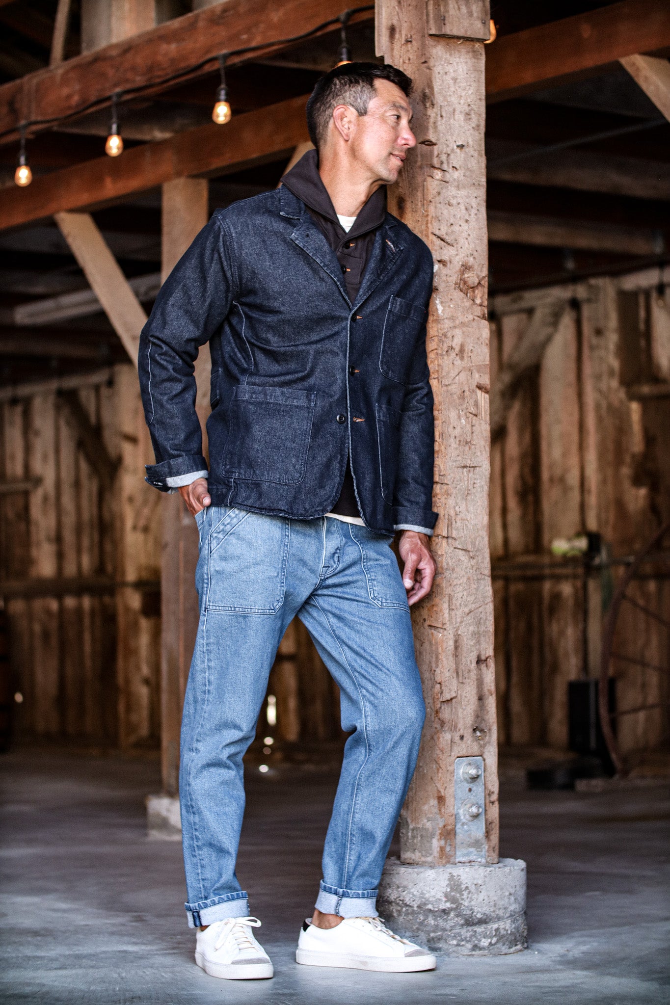 Raw selvedge denim jacket paired with light-wash selvedge denim jeans, styled with a hoodie and white sneakers in a rustic setting.