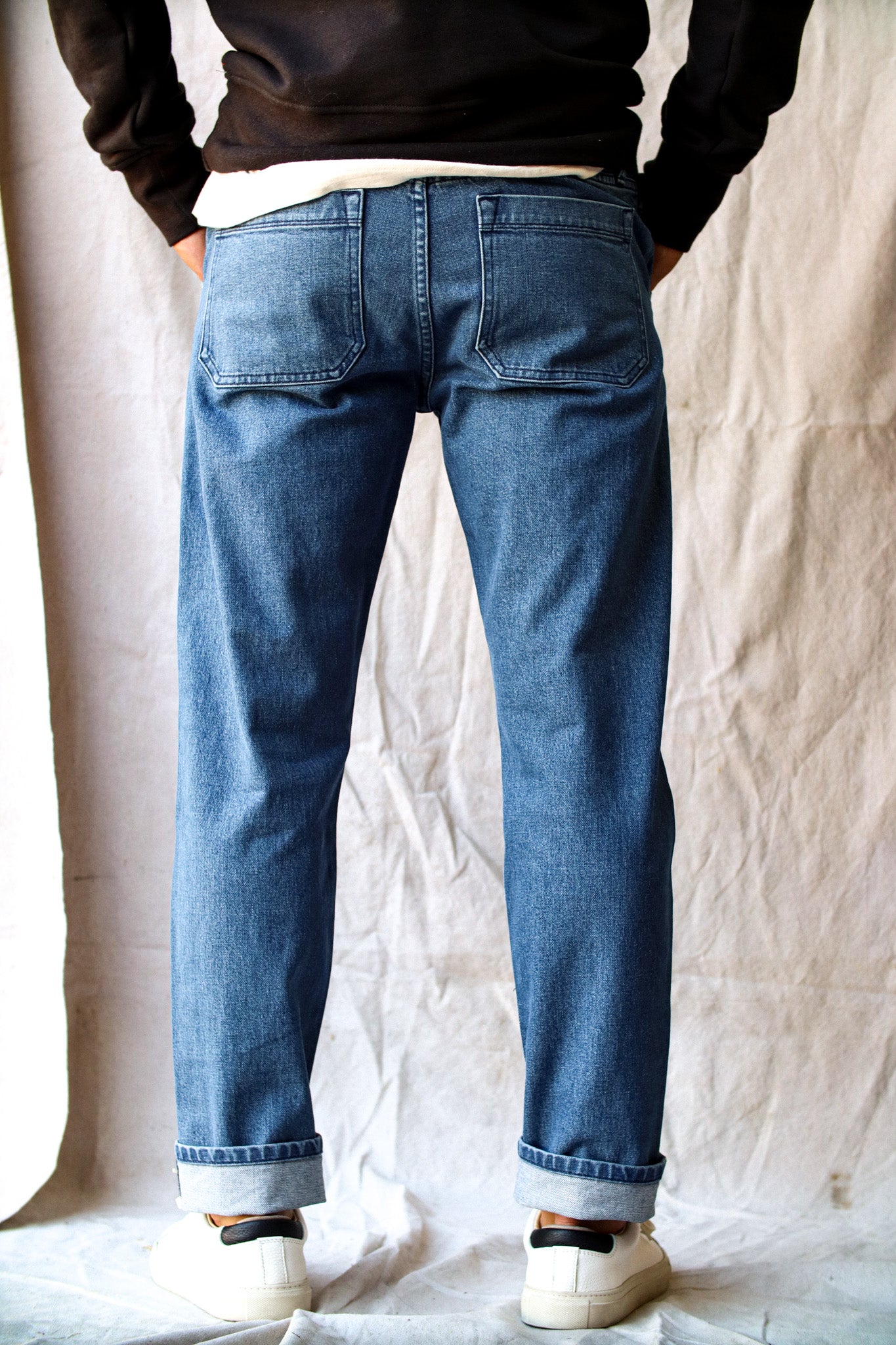 Raw denim jeans with a straight-leg fit, back patch pockets, and cuffed hems, styled with white sneakers and a black sweatshirt.