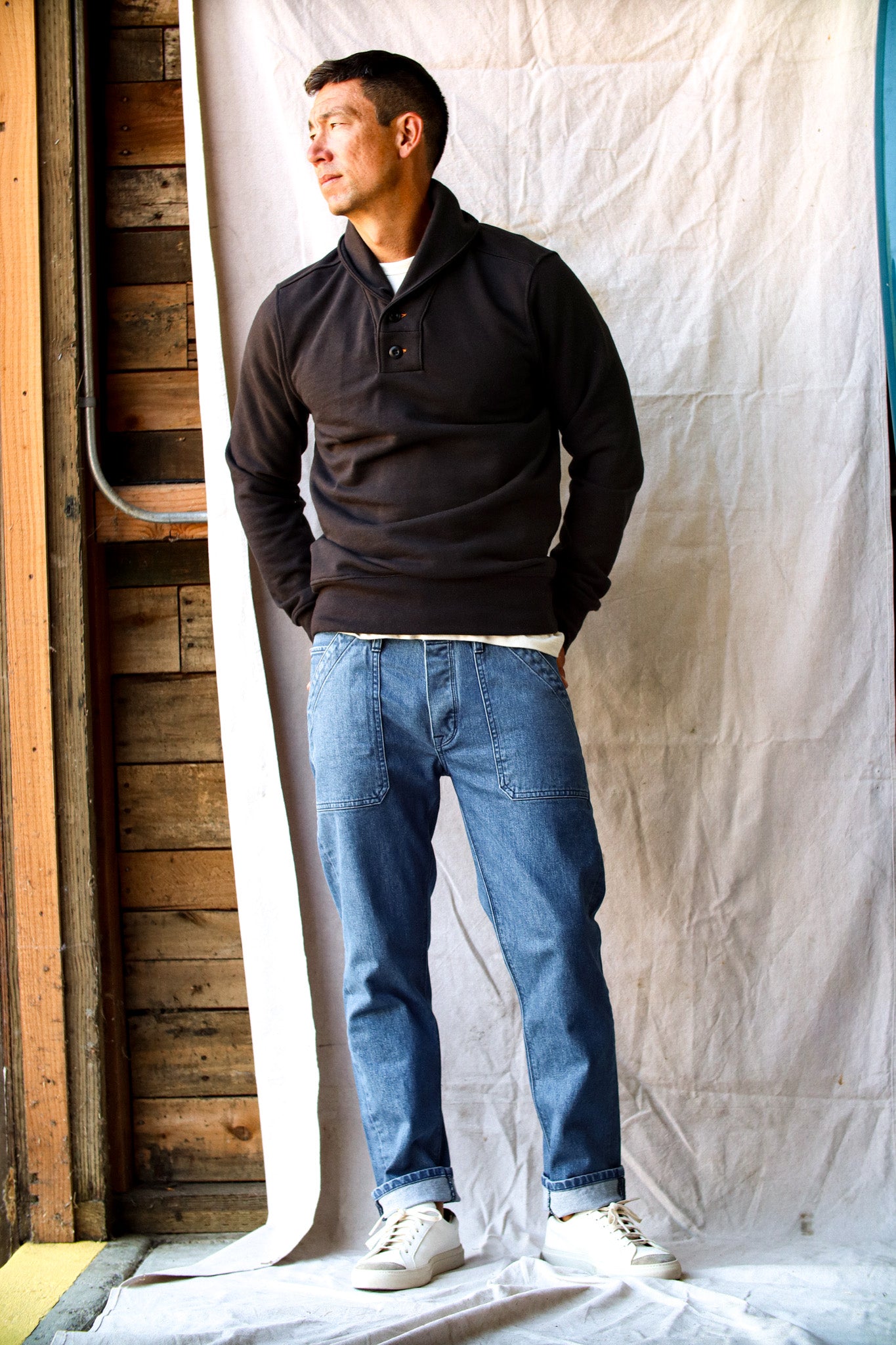 Raw selvedge denim jeans in a relaxed fit, styled with a black pullover and white sneakers against a rustic backdrop.