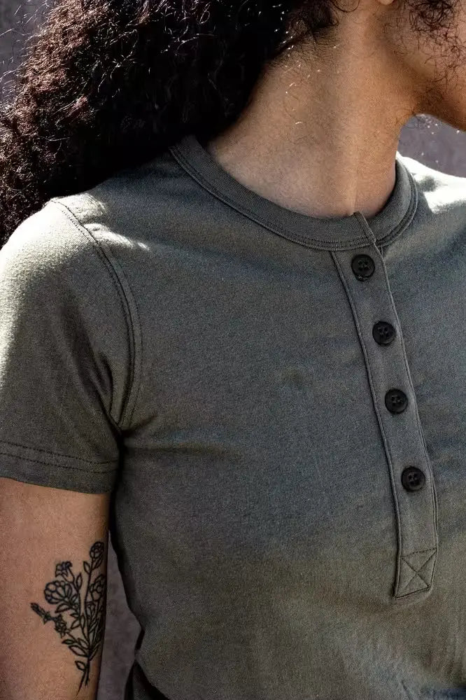 Close-up of a woman wearing the Reyes Henley in olive color, made from heavyweight 8oz 100% cotton, featuring a four-button placket & short sleeves, with a floral tattoo visible on the arm
