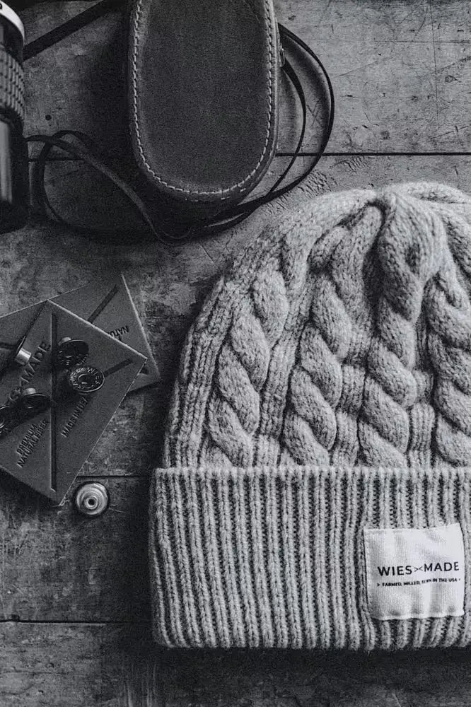 Wool beanie in cable-knit design, navy color, featuring a folded cuff with a branded label, placed on a textured wooden surface alongside leather patches and a leather case