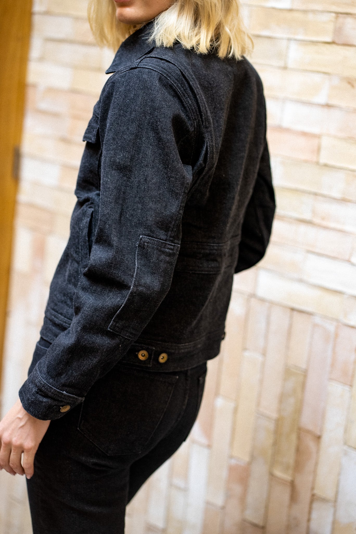 Broken denim jacket worn by a woman with blonde hair, posing against a textured brick wall.