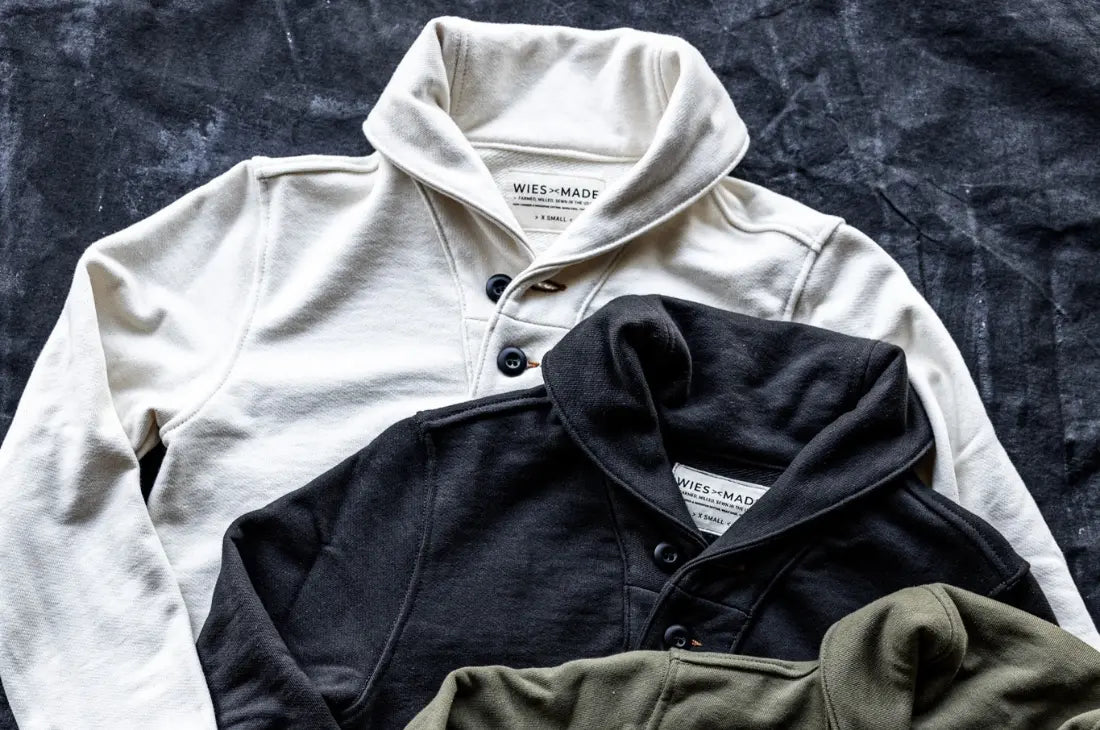 USA-made apparel featuring a collection of high-quality button-up pullover sweaters in cream, black, and olive, laid on a textured surface.