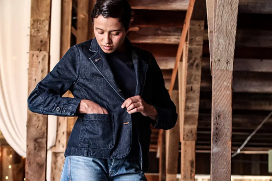 Broken twill denim jacket worn by person adjusting pocket in rustic indoor setting with wooden beams