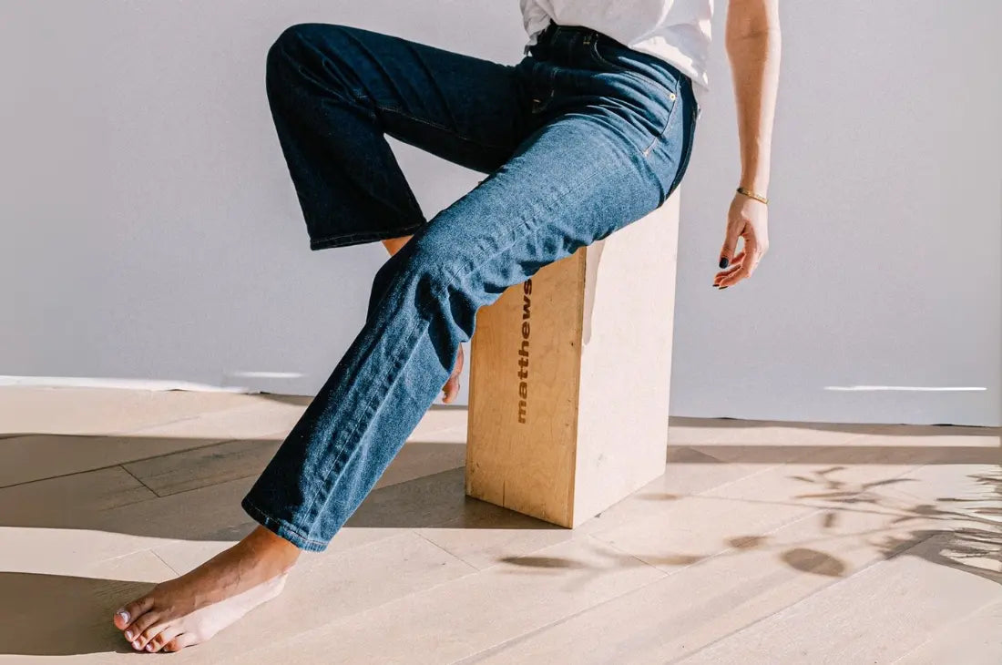 Comfortable washed jeans worn by woman sitting barefoot on wooden block in bright minimalist studio setting