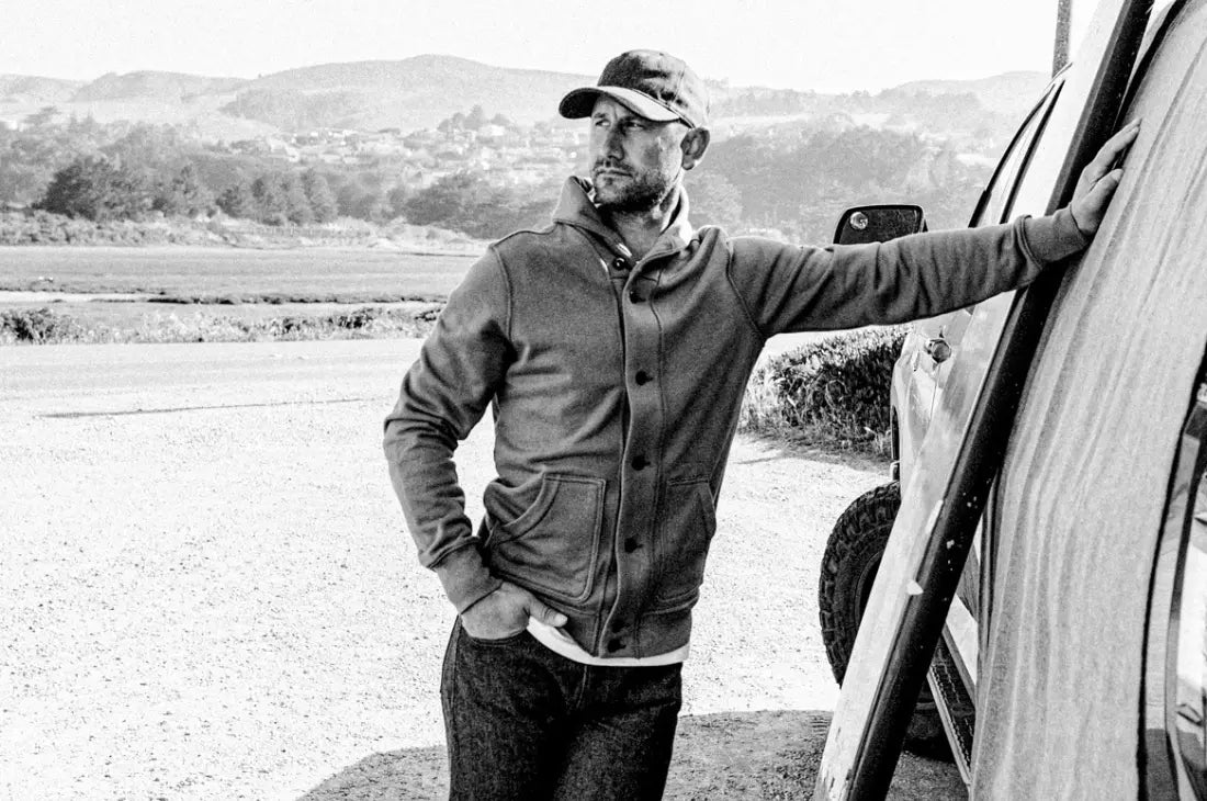 French cardigan worn by man leaning on vehicle in rugged outdoor setting, captured in high-contrast black and white