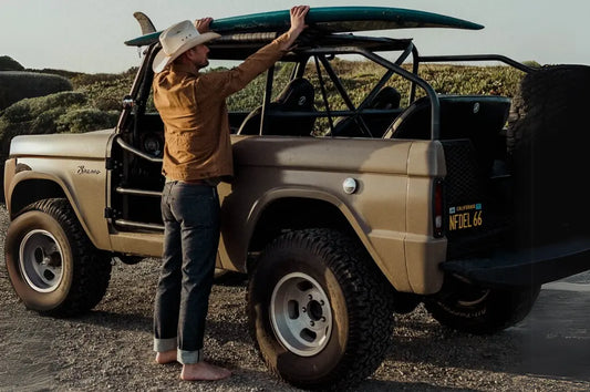 Premium denim jacket paired with cuffed jeans, worn by a man loading a surfboard onto a vintage off-road vehicle