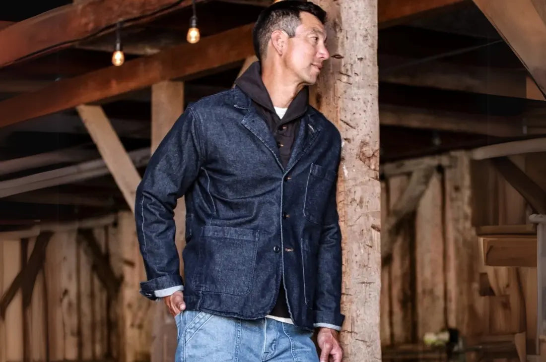 Modern knitwear layered under a denim blazer, worn by a man in a rustic barn setting with warm lighting