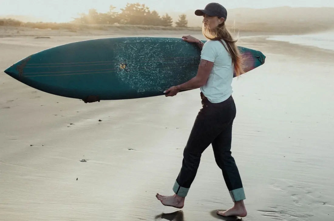 Rigid denim jeans worn by a woman walking barefoot on the beach carrying a surfboard at sunset.