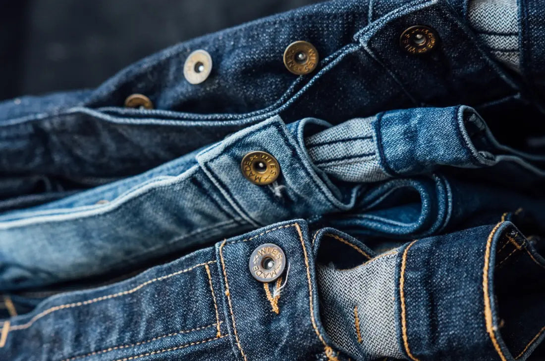 Premium selvedge denim jeans stacked together, showcasing intricate stitching, metal buttons, and high-quality raw denim texture