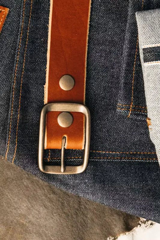 Latigo Belt in 1.5” width, crafted from 8oz American Latigo Leather, featuring a brass buckle placed over raw selvedge denim with visible orange stitching and selvedge edge