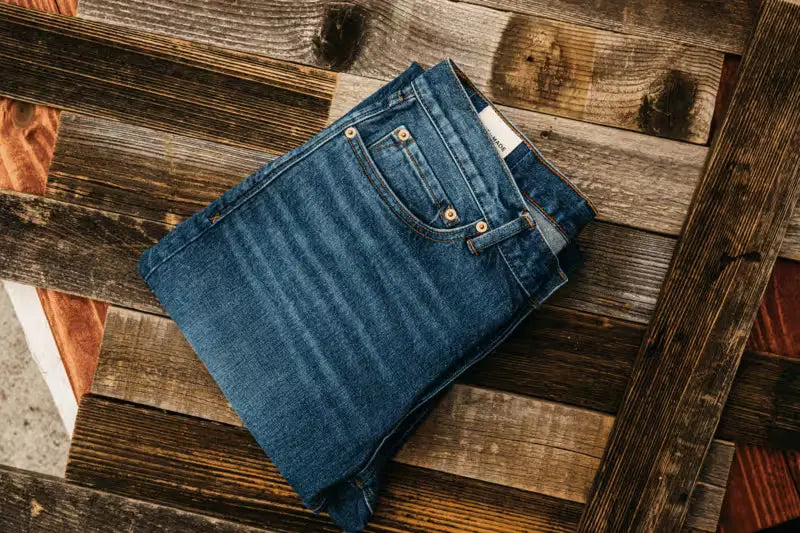 
Sam Fit jeans in River Wash color, neatly folded, showcasing classic stitching and brass rivets, placed on a rustic wooden surface with varying shades of brown and gray plank