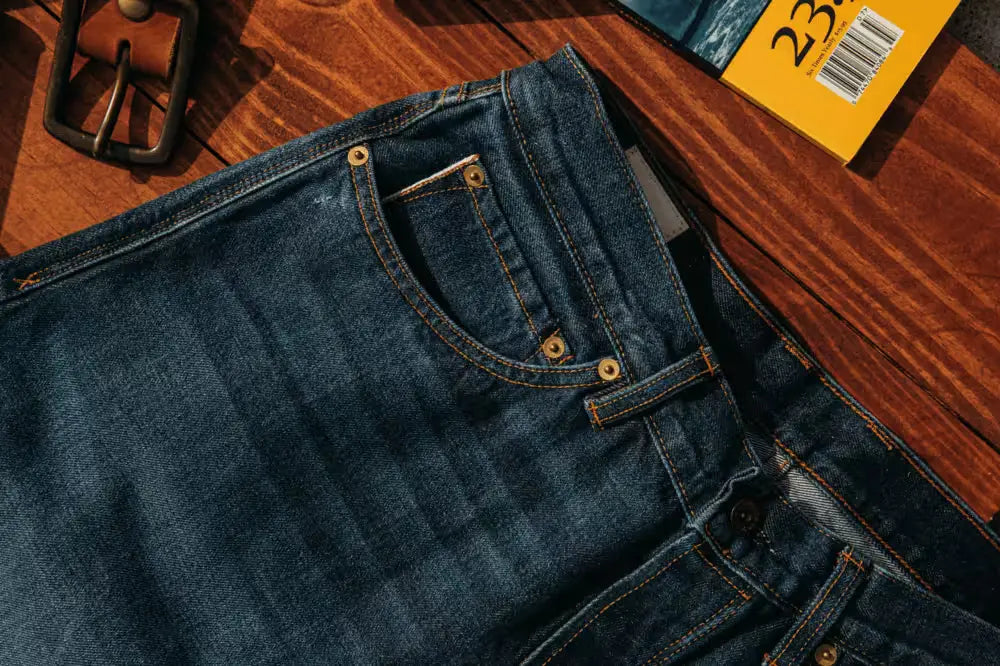 Sam Fit jeans in Washed Selvedge color, showcasing detailed stitching, brass rivets, and a coin pocket, placed on a polished wooden surface with rich grain patterns