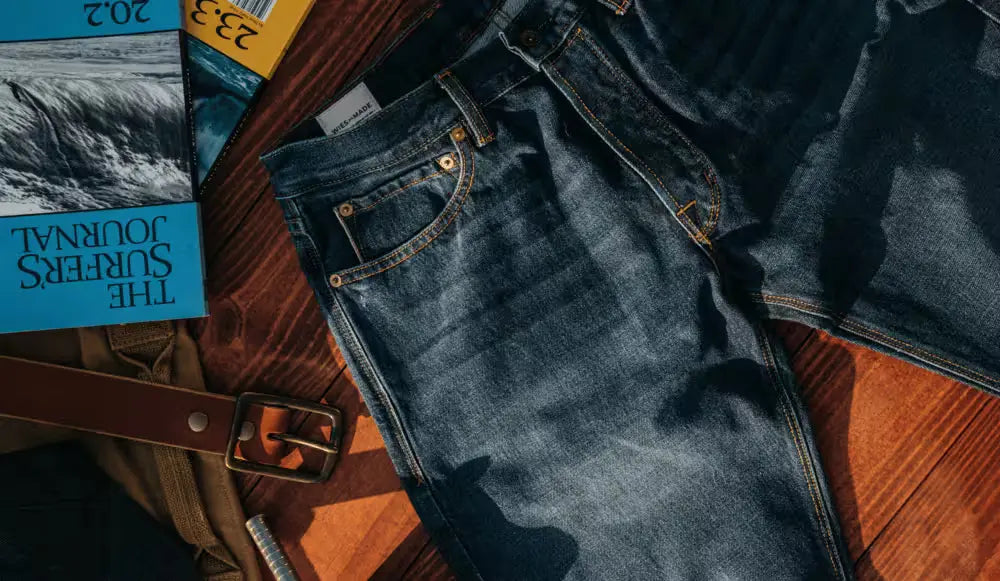 Sam Fit Jean in Ocean Washed Selvedge color, featuring a standard cut, displayed on a wooden surface with a brown leather belt, a metal tool, & surf journals in the background