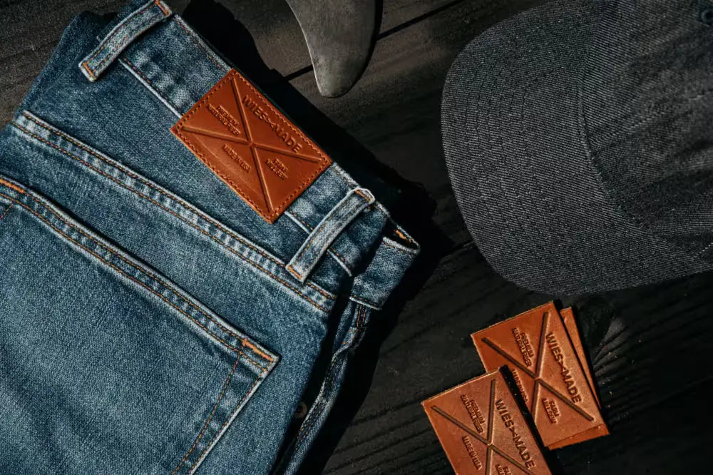 Sam Fit jeans in River Wash color, featuring a leather brand patch and detailed stitching, laid on a dark wooden surface. The background includes additional leather patches with embossed branding, a denim-textured cap, and a metallic tool