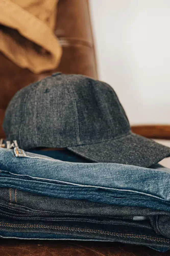 Selvedge denim ballcap resting on a folded stack of denim jeans on a leather chair