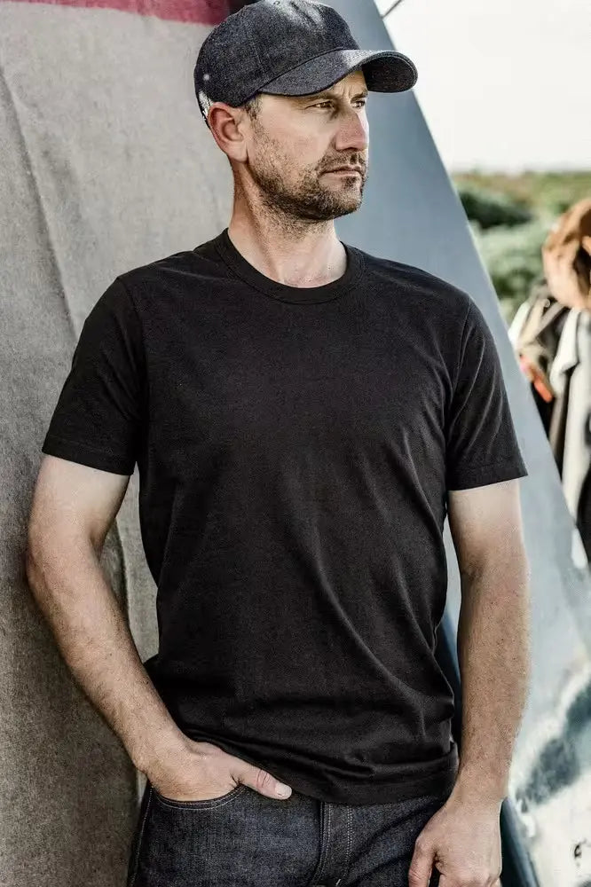 man wearing the Bolinas Tee in basalt color and a cap, made from heavyweight 8oz 100% cotton, standing against a textured backdrop with a surfboard partially visible in the background