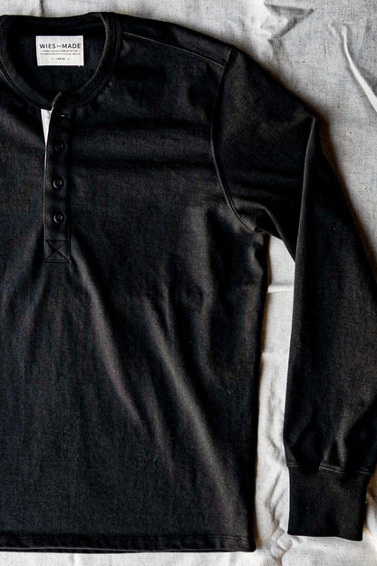 Flat lay detail of a black heavyweight cotton henley on linen showing the button placket and neck label.