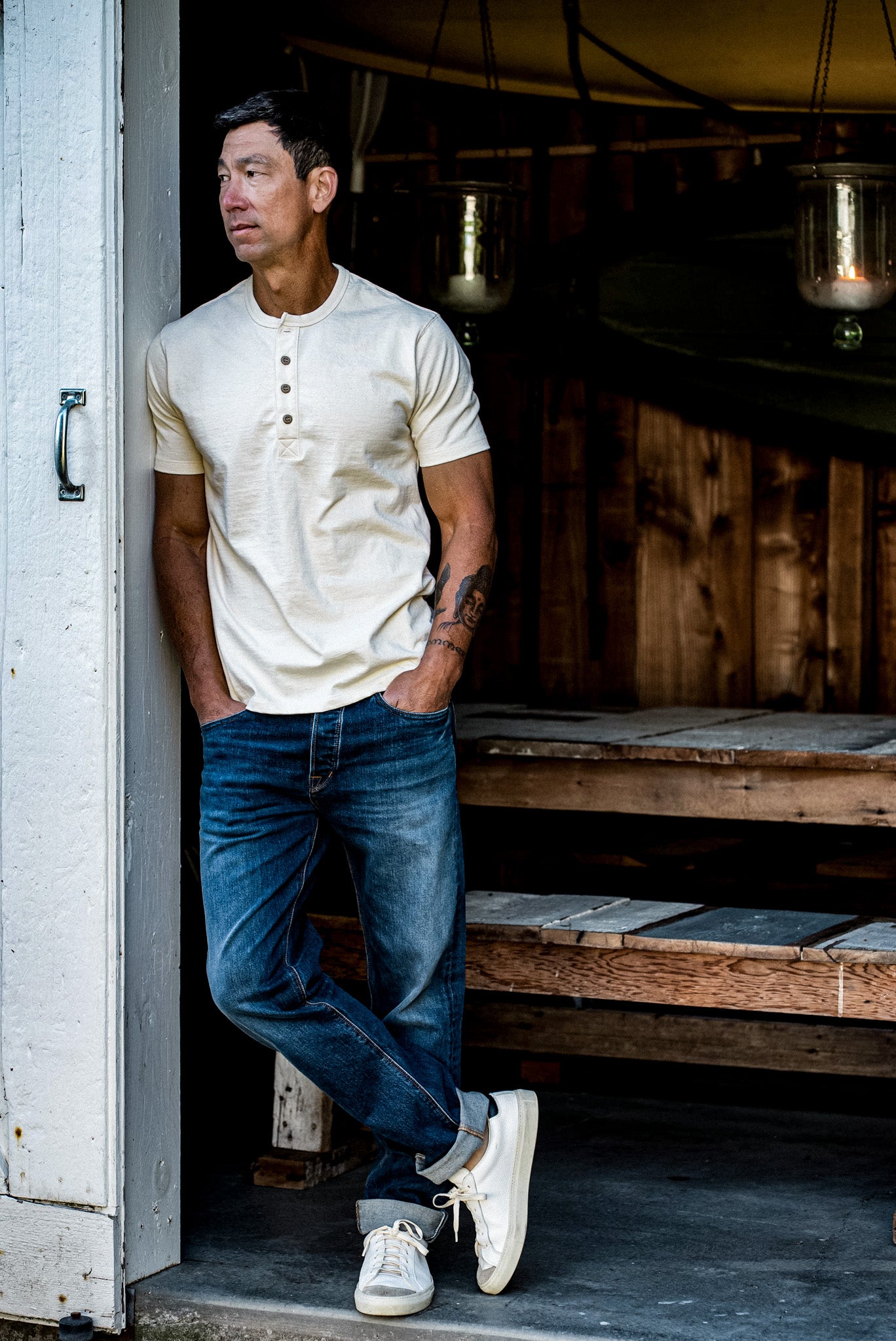 Man in a natural short-sleeve Bolinas henley and cuffed blue jeans leaning in a barn doorway.