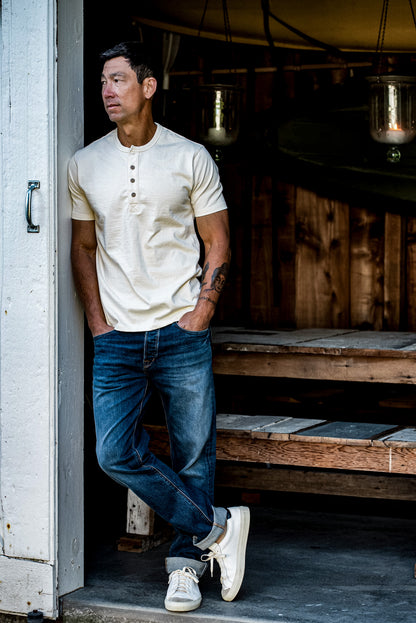Man in a natural short-sleeve Bolinas henley and cuffed blue jeans leaning in a barn doorway.