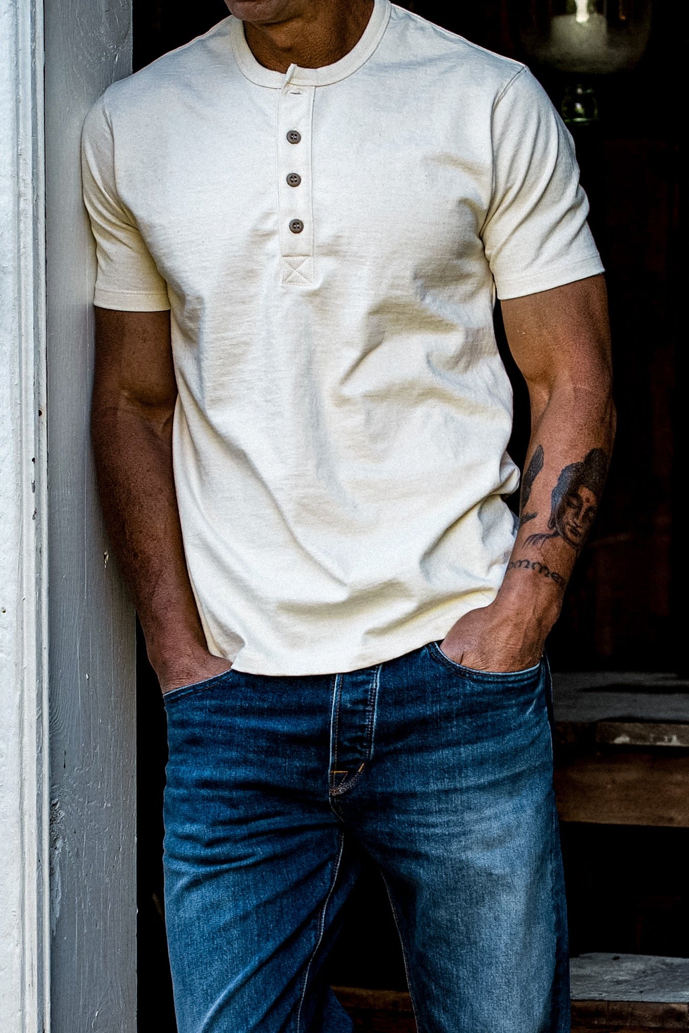 Man in natural cream four-button heavyweight cotton Henley paired with blue denim jeans, leaning in a doorway.